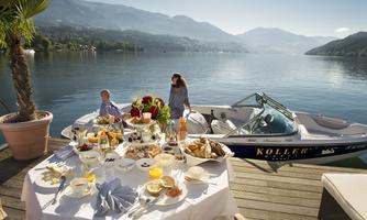 A beautiful table set with delicious food and drinks by the water. In the background, there is a boat and a picturesque mountain landscape.