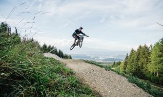A mountain biker jumps over a gentle rise in a green landscape. In the background, you can see trees and a clear sky.