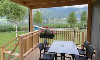 A cozy terrace with a table and chairs, surrounded by green grass and trees. In the background, there is a beautiful lake and mountains visible.