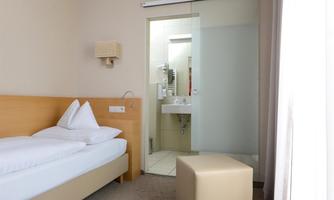 A simple room with a single bed and a modern bathroom. The walls are neutral, and there is a small seating ottoman.