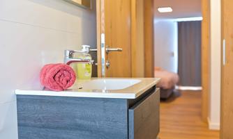 A modern bathroom with a wooden sink and a red towel roll. The background features a bright hallway with a bedroom door.