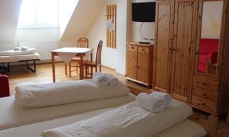 A cozy room with two beds, a table, and a wooden wardrobe. The room has large windows that let in plenty of daylight.