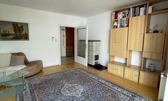 A bright, spacious living area with a large, decorative rug. In the background, there is a bookshelf and an open door leading to the foyer.