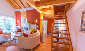 A modern living space with a wooden ceiling and bright walls. A staircase leads to additional rooms, while cozy furniture makes the space inviting.