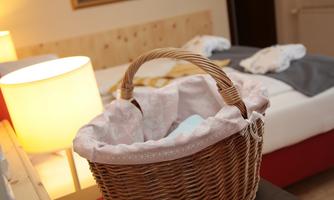 A woven basket stands in a bright room. In the background, a bed and a table lamp can be seen.