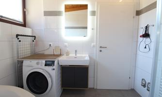 A modern bathroom with a washing machine, a sink, and a large mirror. The walls are tiled and there is plenty of daylight from the window.