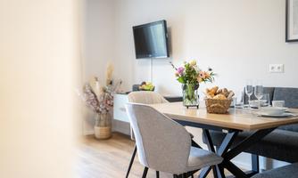 An elegant dining area with a table adorned with flowers and a basket of rolls. In the background, a television is mounted on the wall.