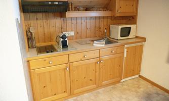 A small kitchen with wooden cabinets, a microwave, and a kettle. The floor is light, and the walls are neutral colors.