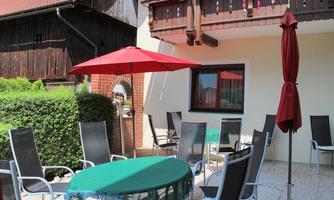 A cozy terrace with a round table and chairs. A red umbrella provides shade and the garden is well maintained.