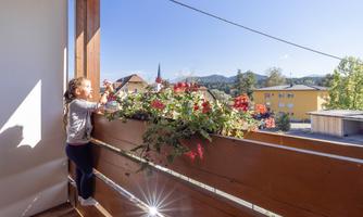 A girl is standing on a balcony watering blooming flowers. In the background, houses and mountains can be seen, while the sun is shining.