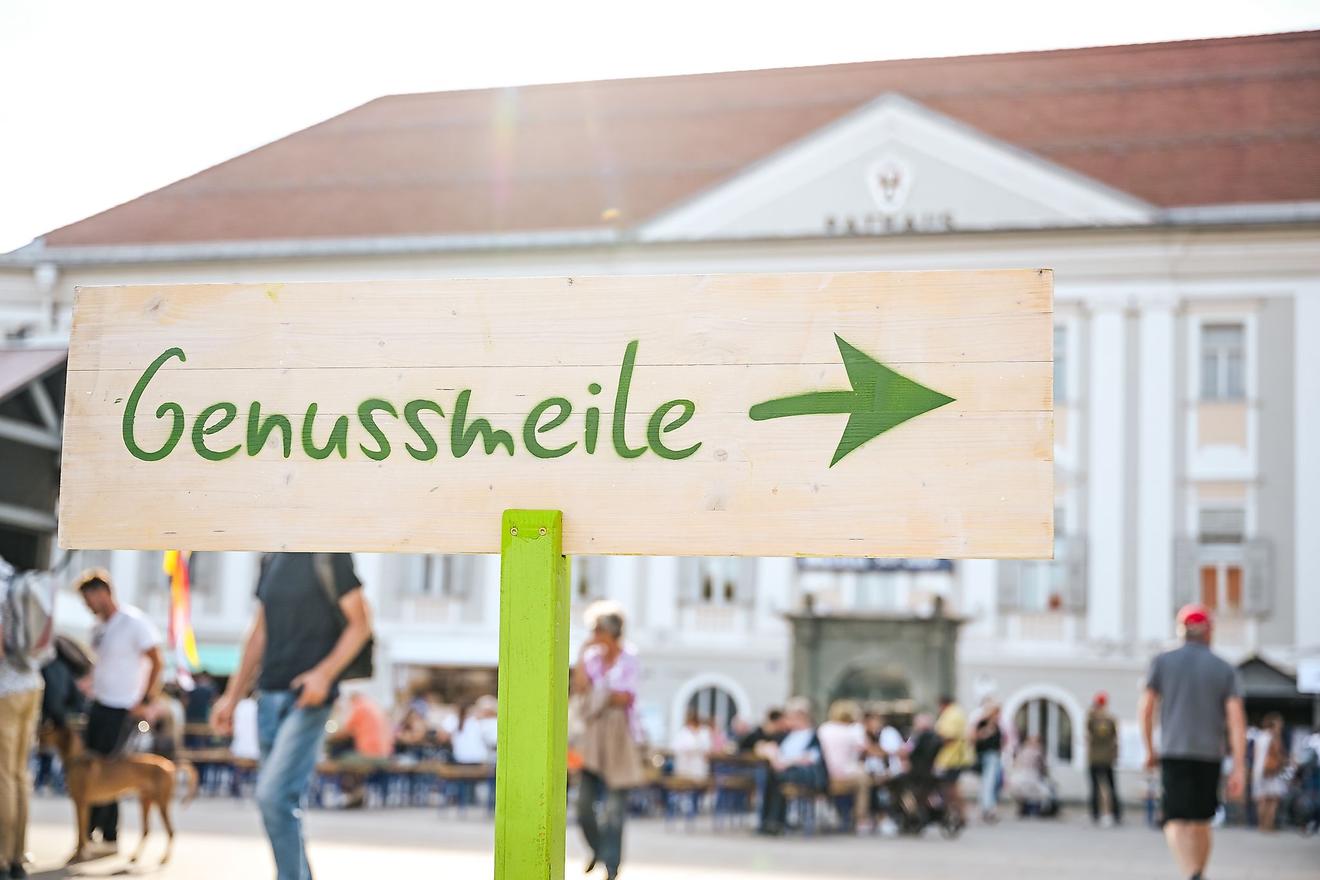 Genussmeile Schild mit grünem Pfeil weist den Weg zu einem Fest mit Menschen