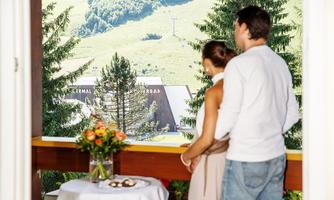 A couple looks from a balcony over a green landscape with trees. On the table, there are flowers and snacks, fitting the romantic atmosphere.