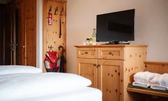 A cozy hotel room with light wooden furniture and a television. A few drinks are on the dresser and a backpack is attached to the wall.