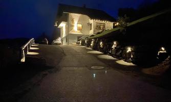 A modern house on a hill at night, illuminated by warm light. The driveway is lined with small lamps and leads to the entrance.