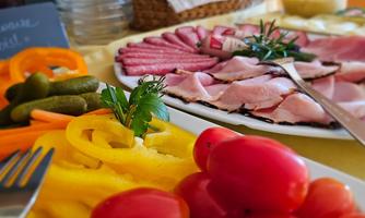 A delicious plate with various types of sausage, fresh vegetables, and cherry tomatoes. In the background, rolls and other snacks can be seen.