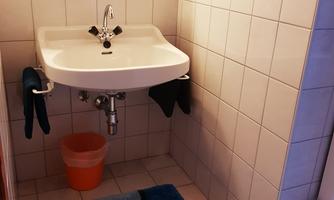 A small bathroom with a sink on the wall. There is a colored rug on the floor and a bucket stands next to it.