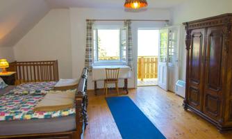 A cozy bedroom with two beds, a table, and a wooden wardrobe. Large windows provide plenty of daylight and access to a balcony.