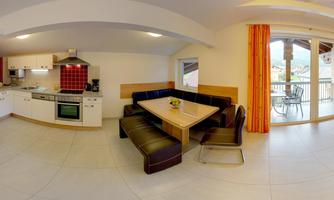 A modern kitchen with a living area and a dining table. Large windows provide views and daylight.