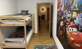 A children's room with a bunk bed and a colorful mural of cars. The room has a light wooden floor and an open door to another room.