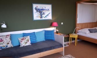 A cozy room with a comfortable sofa and colorful cushions. A colorful picture hangs on the wall, and there is a bed in the corner.