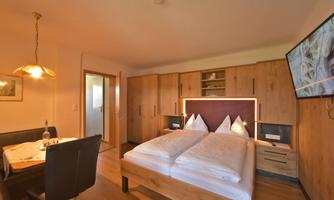 A cozy bedroom with a double bed and wooden furniture. In the corner stands a dining table with chairs and a television on the wall.