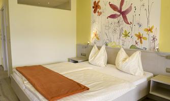 A bright room with a double bed and artistic wall design. On the bed lies an orange blanket and there are two decorative cushions.