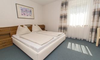 A bright bedroom with a double bed and two pillows. Large windows let in plenty of daylight and the walls are simply decorated.