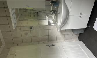 A bright bathroom with a modern sink and a mirror. The walls are tiled and there is a shower in the background.
