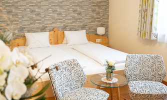 A cozy bedroom with two single beds and decorative cushions. Next to the beds are two stylish chairs and a glass table with a small flower decoration.