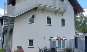 A modern house with multiple windows and a balcony. In the foreground is a terrace with a table and chairs.