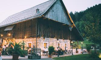 A rustic farmhouse surrounded by trees and plants. People are sitting on the terrace and enjoying the atmosphere.