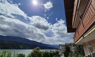 A view of a tranquil lake with mountains in the background and a clear blue sky. Clouds are passing by, and a building is visible in the foreground.
