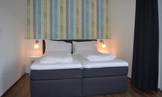 A modern bedroom with a double bed and colorful, striped background. The walls are light and minimalist, with pleasant lighting.