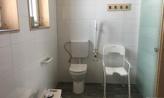 A functional bathroom with a toilet and a chair for additional support. The walls are light and tiled, featuring a modern, clean design.