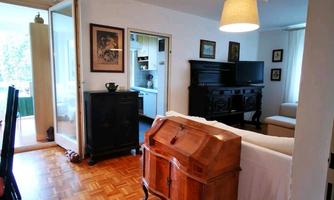 A bright room with wooden floors and a cozy decor. In the foreground, there is an antique piece of furniture, while in the background, another room is visible.