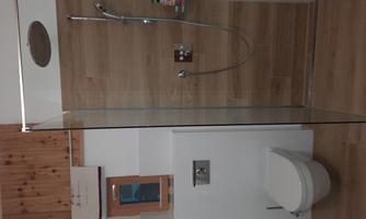 A modern bathroom with a glass shower cabin and wood paneling. The toilet is integrated into the wall, and there is a small window opening.
