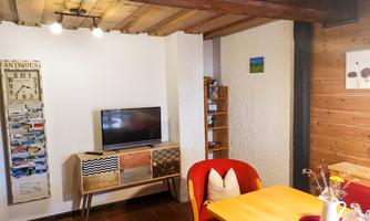 A cozy living ambiance with a wooden table and red chairs. A television and a bookshelf are visible on the wall.