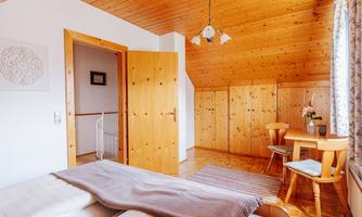 A cozy room with wooden walls and a bright ambiance. There is a double bed, a table with chairs, and a door to another room.