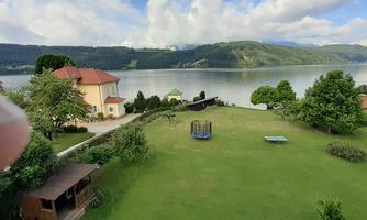 A green meadow by the lake with a lovely garden. In the background, mountains and a clear sky can be seen.