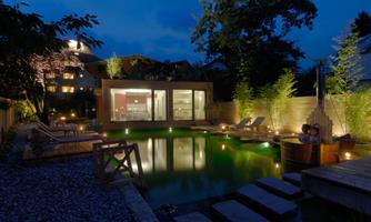 A modern garden with a illuminated pool and wooden decks. In the background, there is a stylish house and it is twilight.
