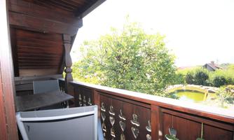 A balcony with a wooden railing, a table, and a chair. In the background, a large tree and a garden can be seen.