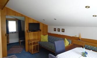 A cozy living room with a blue carpet and wood-paneled walls. There is a table, a television, and a sofa with cushions.