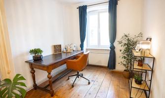 A bright workspace with a large window and modern curtains. There is a wooden desk, a comfortable chair, and some plants in the room.