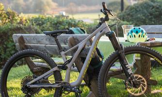 A mountain bike stands next to a wooden bench in a green environment. In the background, trees and a sunny landscape can be seen.
