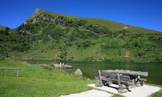A serene landscape with a clear lake and a green meadow. In the foreground, there is a wooden bench under a blue sky.