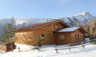A cozy wooden house in the snowy landscape. In the background, mountains and a clear sky can be seen.