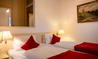 A cozy bedroom with two beds and red pillows. A landscape painting hangs on the wall, and warm lamps create an inviting atmosphere.