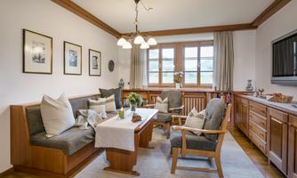 A cozy living room with wooden decor, comfortable chairs, and a bright window front. Fresh flowers are on the table, and pictures are hanging on the wall.