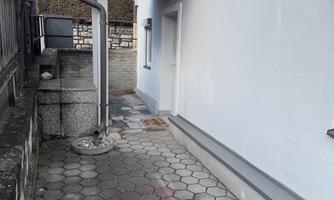 A narrow path along a house wall with gray paving stones. Above the entrance, there is a small wooden awning.