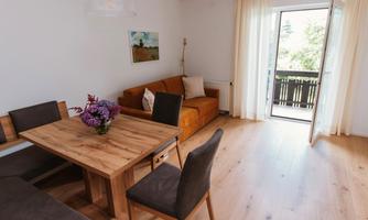 A bright living room with a wooden dining table and a cozy sofa. Large windows offer a view of the balcony and the outdoor area.